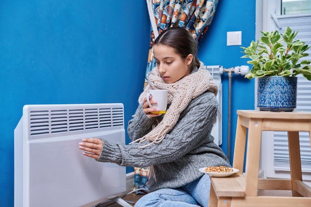 Autunno freddo stagione invernale stile di vita adolescente femminile in maglione caldo sciarpa riscaldamento vicino riscaldamento elettrico radiatore bere bevande calde con tazza apparecchiature di riscaldamento calore vita nella stagione fredda