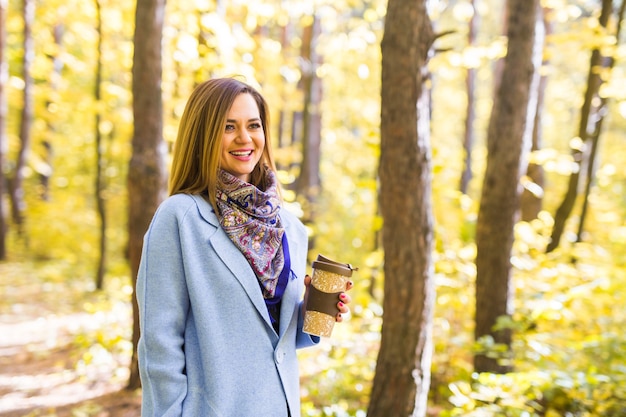 Autunno, natura, concetto di persone - giovane donna bruna in un cappotto blu in piedi nel parco con una tazza