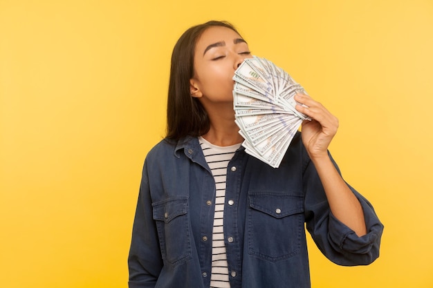 Avido di soldi. Ritratto di avida ragazza ricca in camicia di jeans che odora di banconote in dollari guadagnate, godendo del successo e del grande profitto, vita ricca. tiro in studio indoor isolato su sfondo giallo