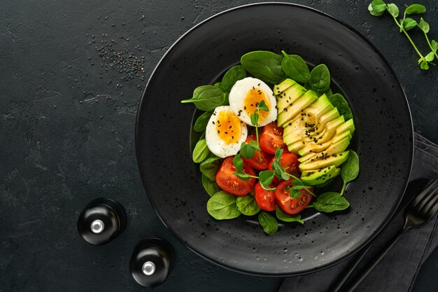 Avocado, pomodorini, spinaci e uova di gallina, piselli microgreens e semi di sesamo nero insalata fresca in una ciotola nera su sfondo nero di ardesia, pietra o cemento. Concetto di cibo sano. Vista dall'alto.