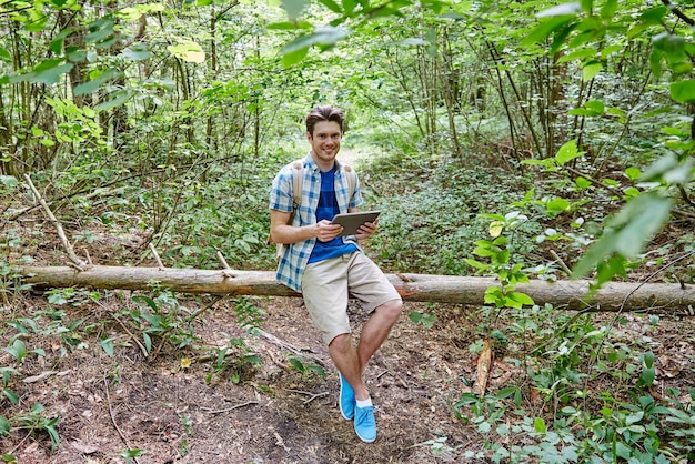 avventura, viaggi, turismo, escursione e concetto di persone - giovane felice con zaino e computer tablet pc seduto sul tronco d'albero caduto nei boschi
