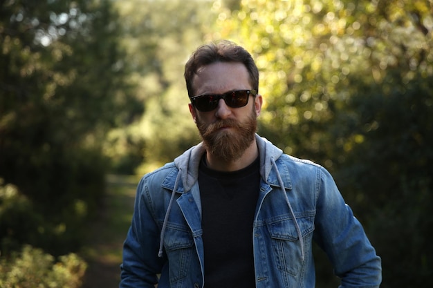 Avventuriero barbuto uomo caucasico con giacca di jeans e occhiali da sole in posa in natura