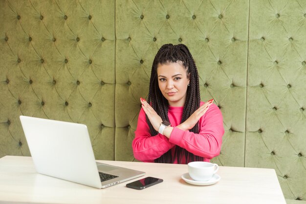 Avvertimento ragazza aggressiva con dreadlocks neri in camicetta rosa sono seduti al bar e lavorando su un laptop che mostra incrociare le braccia alzate come stop o fine gesto guardando la fotocamera interna