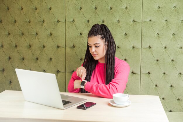 Avvertimento seria giovane donna d'affari prepotente con dreadlock neri, seduta e fare videochiamate sul laptop e mostrare il tempo di controllo, puntare il dito verso l'orologio intelligente, ripresa in studio al coperto.