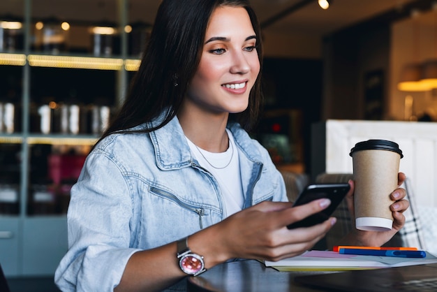 Avvicinamento. Bella sorridente giovane donna d'affari si siede davanti al computer portatile, tiene la tazza di caffè, utilizza lo smartphone per il lavoro