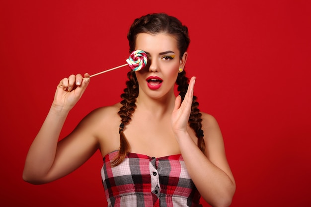 Avvicinamento. Ragazza bruna con labbra rosse, tenendo lecca-lecca colorata. Le caramelle dolci del ritratto di modello di bellezza su una parete rossa.