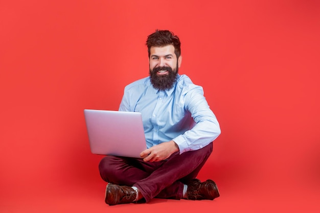 Azienda computer portatile Sorridente bel portatile uomo barbuto lavoratore Felice giovane seduto sul pavimento con e utilizzando il computer portatile su sfondo rosso