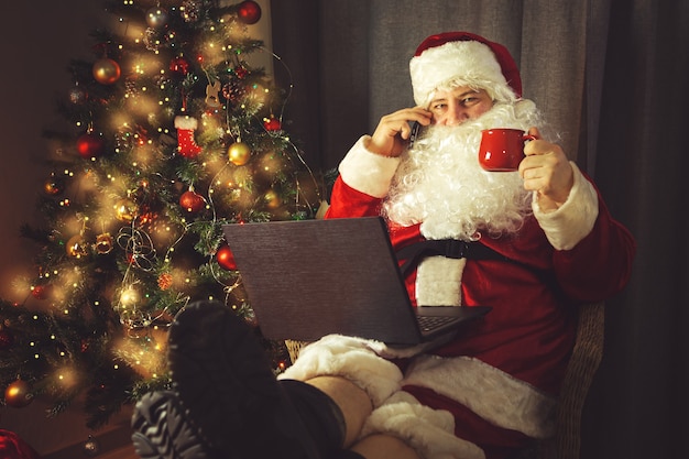 Babbo Natale autentico sta lavorando su un laptop e sta parlando al telefono. Interno di casa. In attesa di Natale e Capodanno.