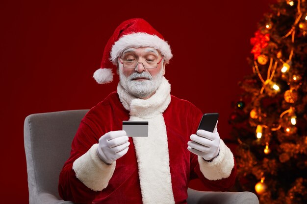 Babbo Natale barbuto in occhiali tenendo la carta di credito e pagando online utilizzando il suo telefono cellulare