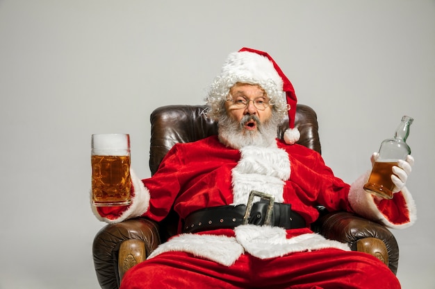 Babbo Natale beve birra seduto sulla poltrona, congratulandosi, sembra ubriaco e felice. Modello maschio caucasico in costume tradizionale. Capodanno 2020, regali, vacanze, atmosfera invernale. Copyspace per il tuo annuncio.