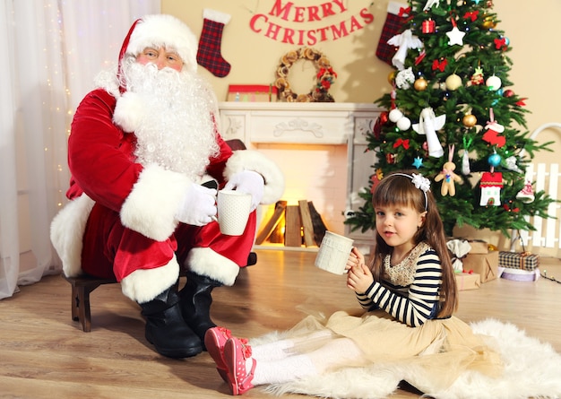 Babbo Natale che beve cioccolata calda con una bambina carina vicino all'albero di Natale a casa