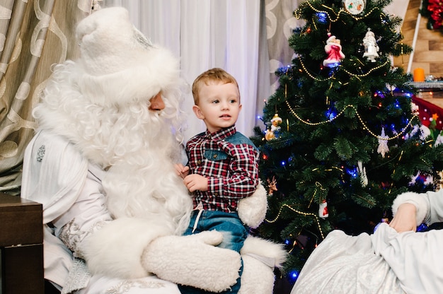 Babbo Natale che fa un regalo a un ragazzino carino in grembo vicino all'albero di Natale a casa.