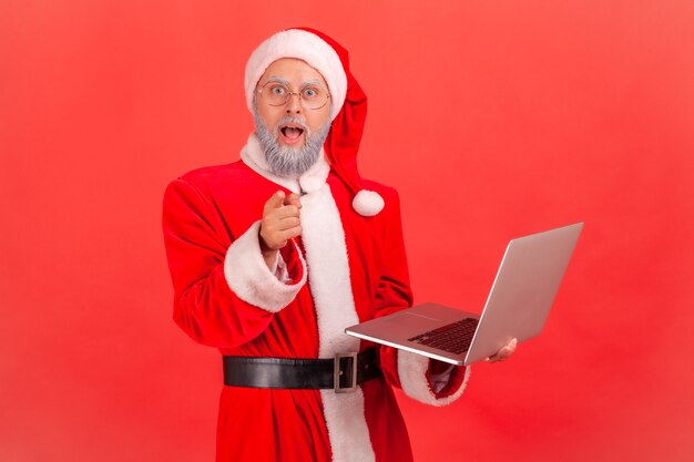 Babbo natale che tiene il portatile in mano e punta alla telecamera con grandi occhi e stupore.