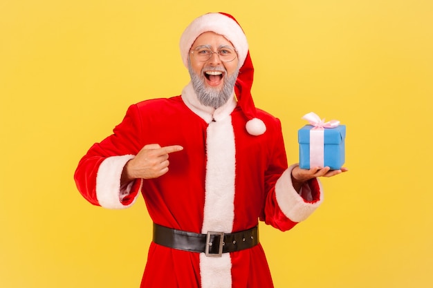 Babbo natale che tiene in mano una scatola e indica un regalo con uno sguardo positivo.