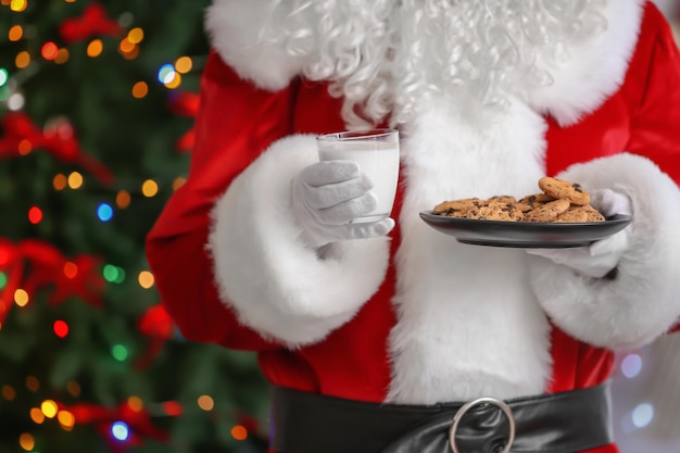 Babbo Natale che tiene piatto con biscotti e bicchiere di latte contro le luci di Natale sfocate