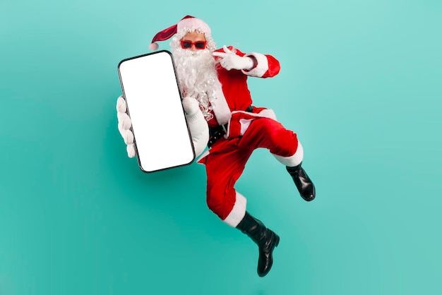 Babbo Natale con gli occhiali da sole e il vestito salta e vola e mostra lo schermo vuoto dello smartphone