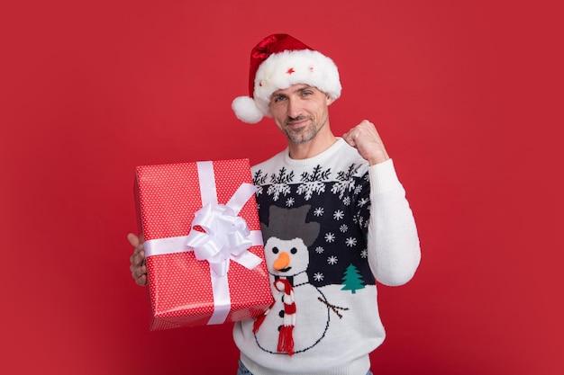 Babbo Natale con il segno del vincitore tiene il regalo sullo sfondo rosso dello studio felice giovane uomo di colore con cappello e maglione di Babbo Natale o Capodanno