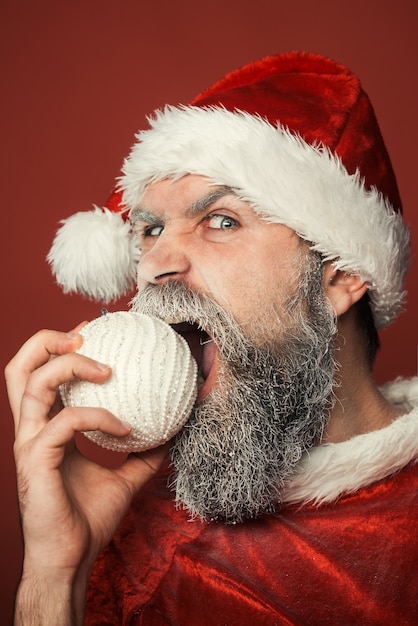 Babbo natale con la barba congelata che mangia il giocattolo decorativo dell'albero di natale su sfondo rosso capodanno e