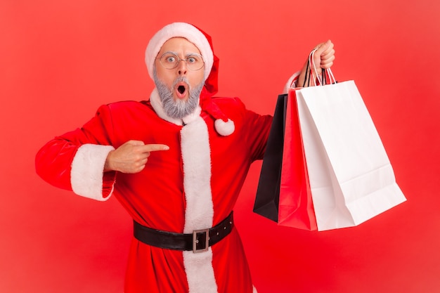 Babbo Natale in piedi che indica le borse della spesa, impressionato dall'acquisto di un acquisto a prezzi bassi.