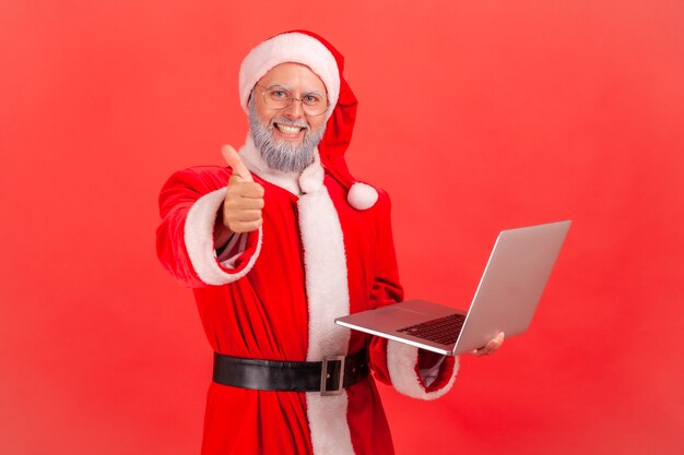 Babbo Natale in piedi con espressione positiva, mostrando pollice in su, tenendo il computer.