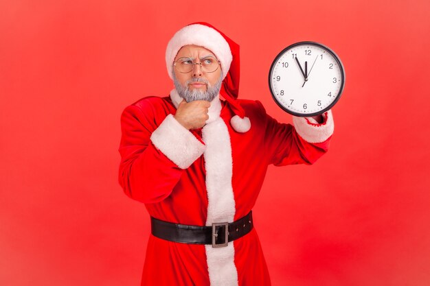 Babbo Natale in piedi con orologio da parete, tenendo il mento, aspettando la festa di Natale, guardando lontano.