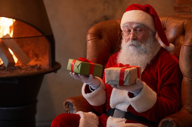 Babbo Natale pensieroso seduto su una comoda poltrona al caminetto e confrontando i regali di natale