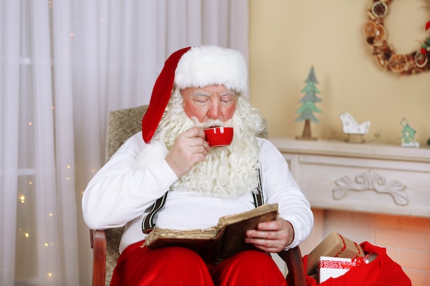 Babbo Natale seduto con il libro in una comoda sedia vicino al camino a casa