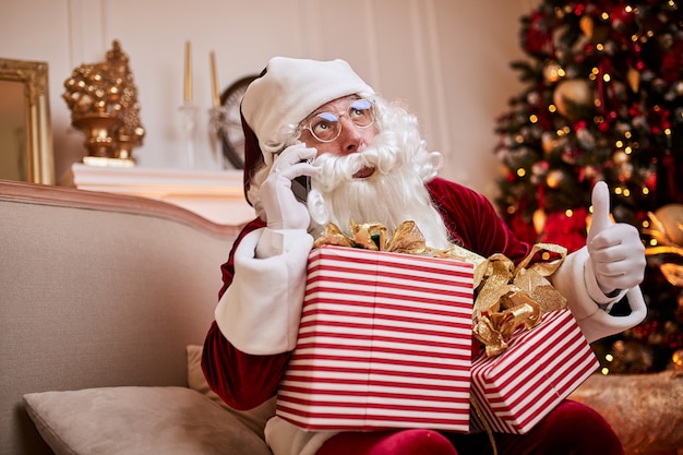 Babbo Natale seduto sul divano e parlando al cellulare vicino al camino e albero di Natale con doni. Buon Natale, concetto di buone feste