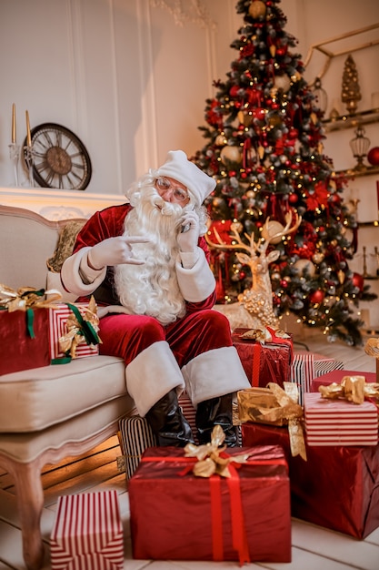 Babbo Natale seduto sul divano e parlando al cellulare vicino al camino e all'albero di natale con i regali. Anno nuovo e buon Natale, concetto di buone feste