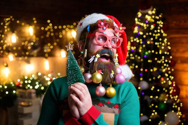 Babbo Natale sorpreso con palle di capodanno in barba tiene Babbo Natale barbuto di abete in occhiali da festa