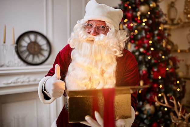 Babbo Natale sorpreso con un magico regalo luminoso che mostra il pollice in su vicino al bellissimo albero di Natale. Anno nuovo e buon Natale, concetto di buone feste