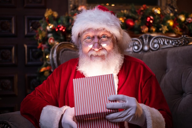 Babbo Natale sorridente tiene il regalo di Natale che soddisfa i desideri di Babbo Natale seduto a casa vicino a Natale