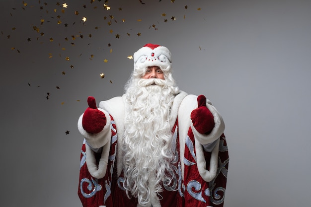 Babbo Natale sotto coriandoli dorati in posa con il pollice in alto sullo sfondo grigio dello studio, Natale festivo e emozioni di capodanno