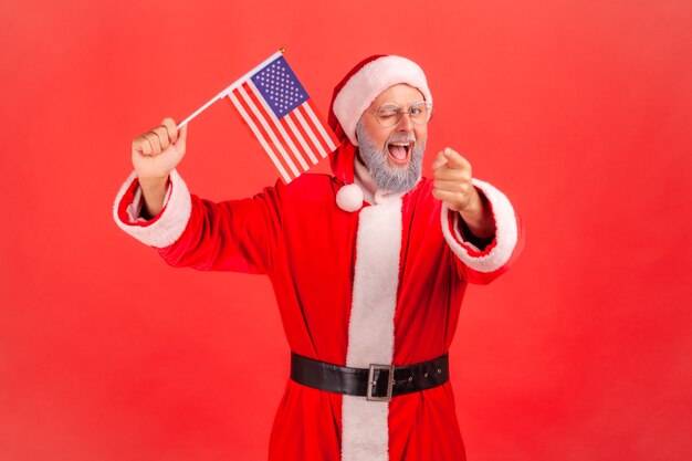 Babbo natale sventola bandiera USA strizzando l'occhio e puntando alla telecamera, tiene la bocca aperta.