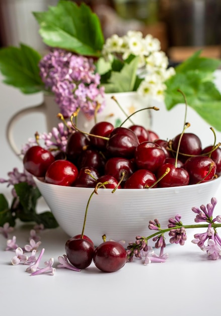 Bacche di succose ciliegie mature in una tazza bianca su uno sfondo di lillà e fiori bianchi