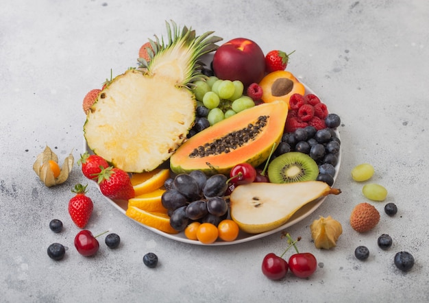 Bacche estive organiche crude fresche e frutti esotici in piatto bianco su sfondo chiaro. Ananas, papaia, uva, nettarina, arancia, albicocca, kiwi, pera, litchi, ciliegia e physalis. Vista dall'alto