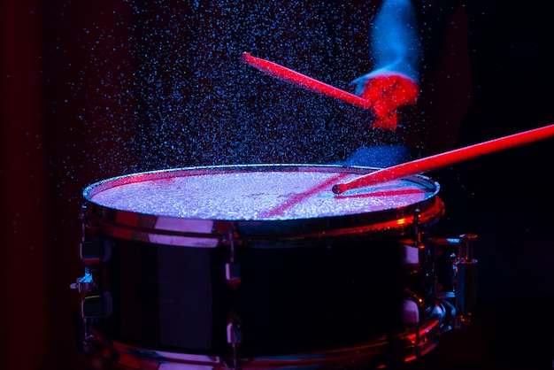 Bacchette di tamburo che colpiscono il rullante con spruzzi d'acqua su sfondo scuro con luci da studio rosse e blu