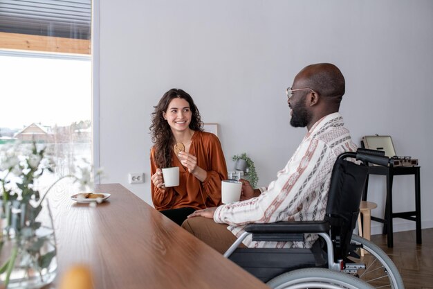 Badante femminile con una tazza di tè e un biscotto che guarda l'uomo di colore con disabilità