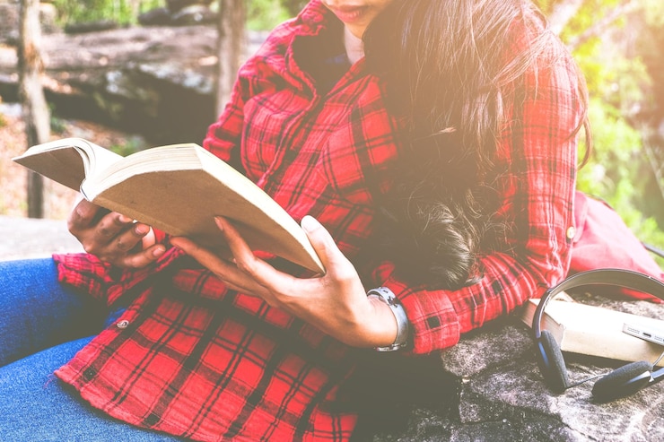 Photo bagian tengah tubuh seorang wanita membaca buku sambil bersandar pada batu