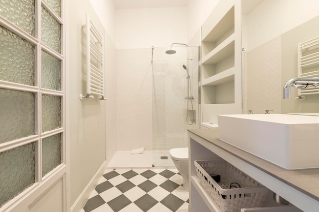 Bagno con mobile da incasso bianco e grigio, cabina doccia lavello in porcellana con divisorio in vetro e pavimento piastrellato a scacchi