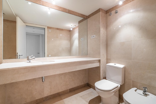 Bagno moderno e spazioso con piastrelle beige luminose, bidet e lavandino su un lungo piano di lavoro in marmo