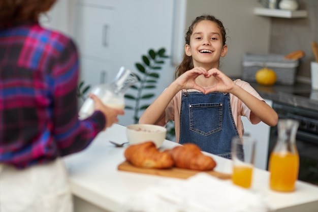 Bambina abbastanza riccia che indossa jeans e maglietta in cucina con sua madre, guardando la telecamera sorridendo facendo la forma del simbolo del cuore con le mani, fa colazione insieme.