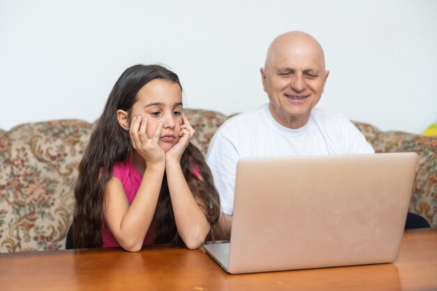 Bambina adorabile che abbraccia nonno felice usando il computer portatile a casa.