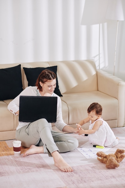 Bambina adorabile che dà la penna a sua madre che è seduta sul pavimento in soggiorno e lavora al computer portatile e riempie i documenti