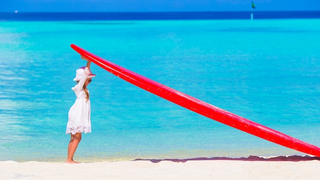 Bambina adorabile con il grande surf rosso sulla spiaggia bianca tropicale