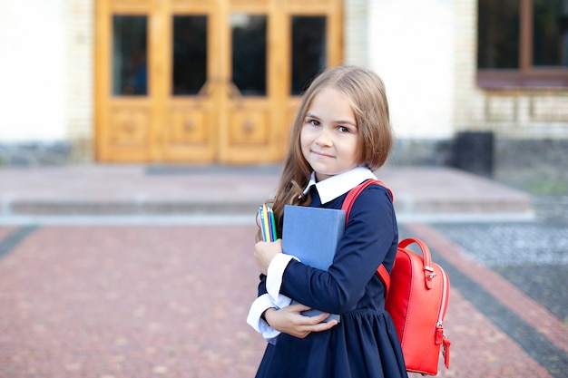 Bambina all'aperto vicino all'edificio scolastico. Studentessa con un libro, una matita e un sacchetto di scuola.