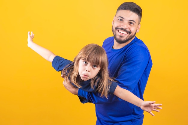 Bambina allegra che gioca con suo padre su sfondo giallo allungando le mani fingendo di volare Giovane padre che tiene sua figlia divertendosi insieme