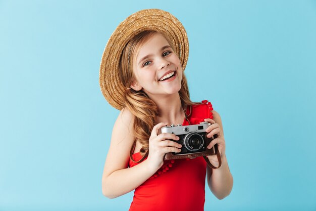 Bambina allegra che indossa il costume da bagno e il cappello estivo in piedi isolato sul muro blu, scattando foto con la macchina fotografica