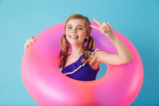 Bambina allegra che indossa il costume da bagno in piedi isolata sul muro blu, giocando con un anello gonfiabile