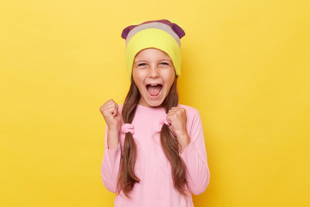 Bambina allegra estremamente felice con le code di cavallo che indossa camicia rosa e cappello a cuffia in posa isolata su sfondo giallo pugno chiuso esultando per il suo successo